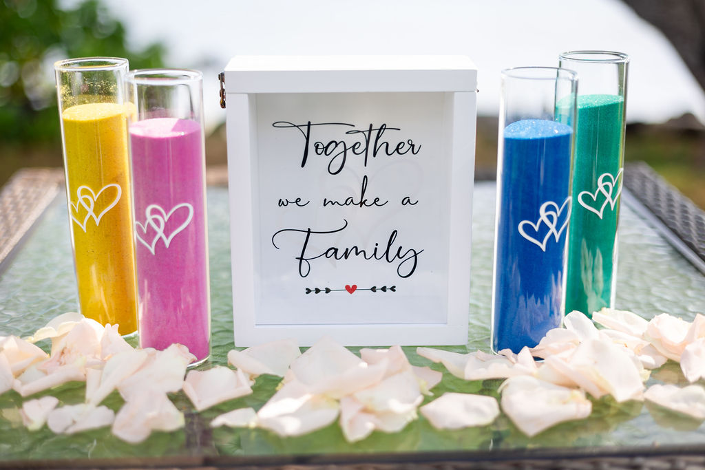 A vibrant family unity sand ceremony setup for an Oahu elopement. Four glass cylinders filled with yellow, pink, blue, and teal craft sand stand next to a white shadow box that reads "Together we make a Family." The arrangement is set on a glass table adorned with white rose petals, overlooking a soft-focus Hawaiian coastal background.