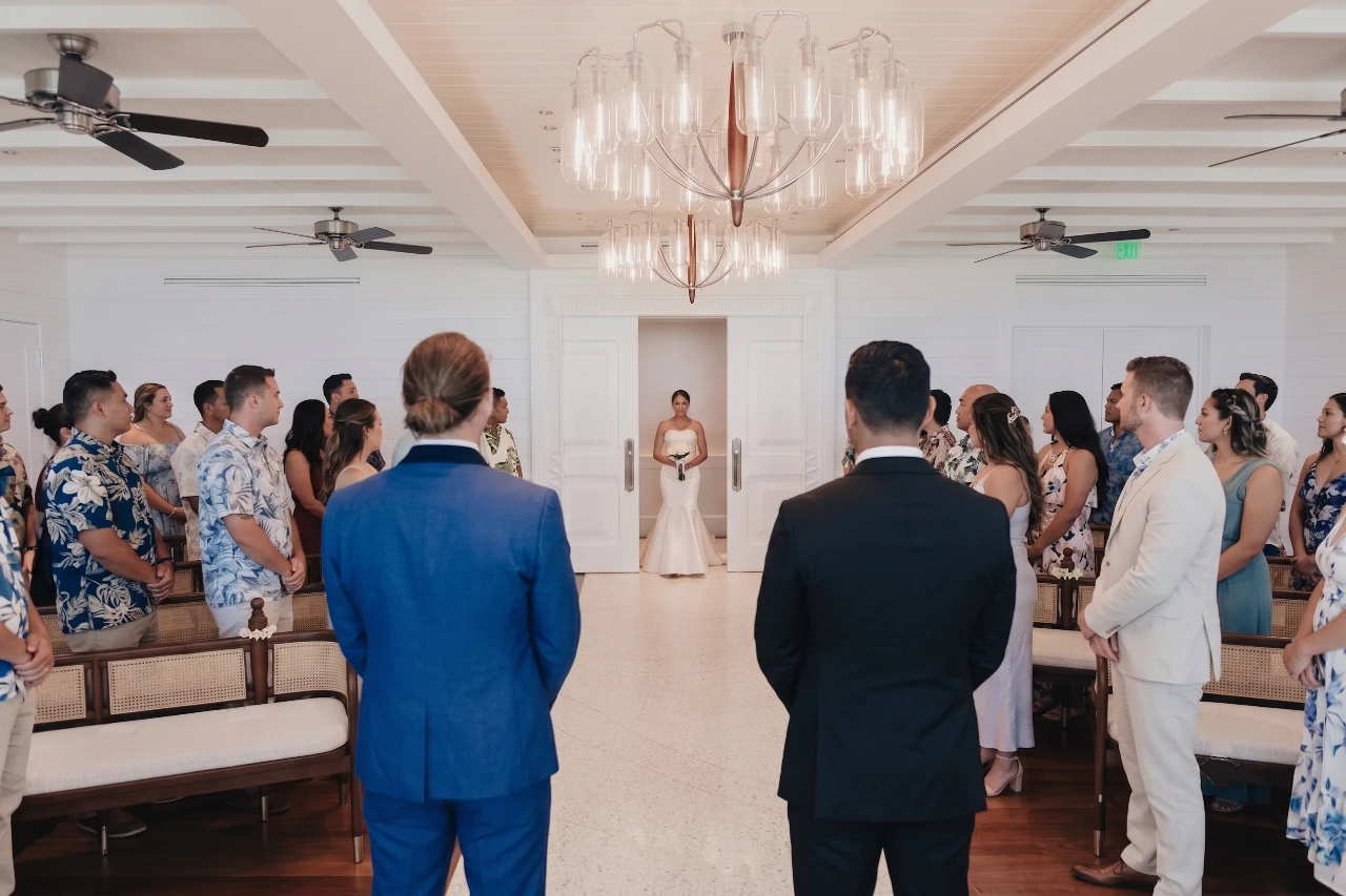 A view from the back of the chapel aisle looking toward a bride standing in an open doorway wearing a strapless mermaid-style wedding gown. Guests stand on both sides in the pews, looking toward her, with a man in a blue suit on the left and a man in a black suit on the right standing in the foreground, facing the bride under elegant glass chandeliers.