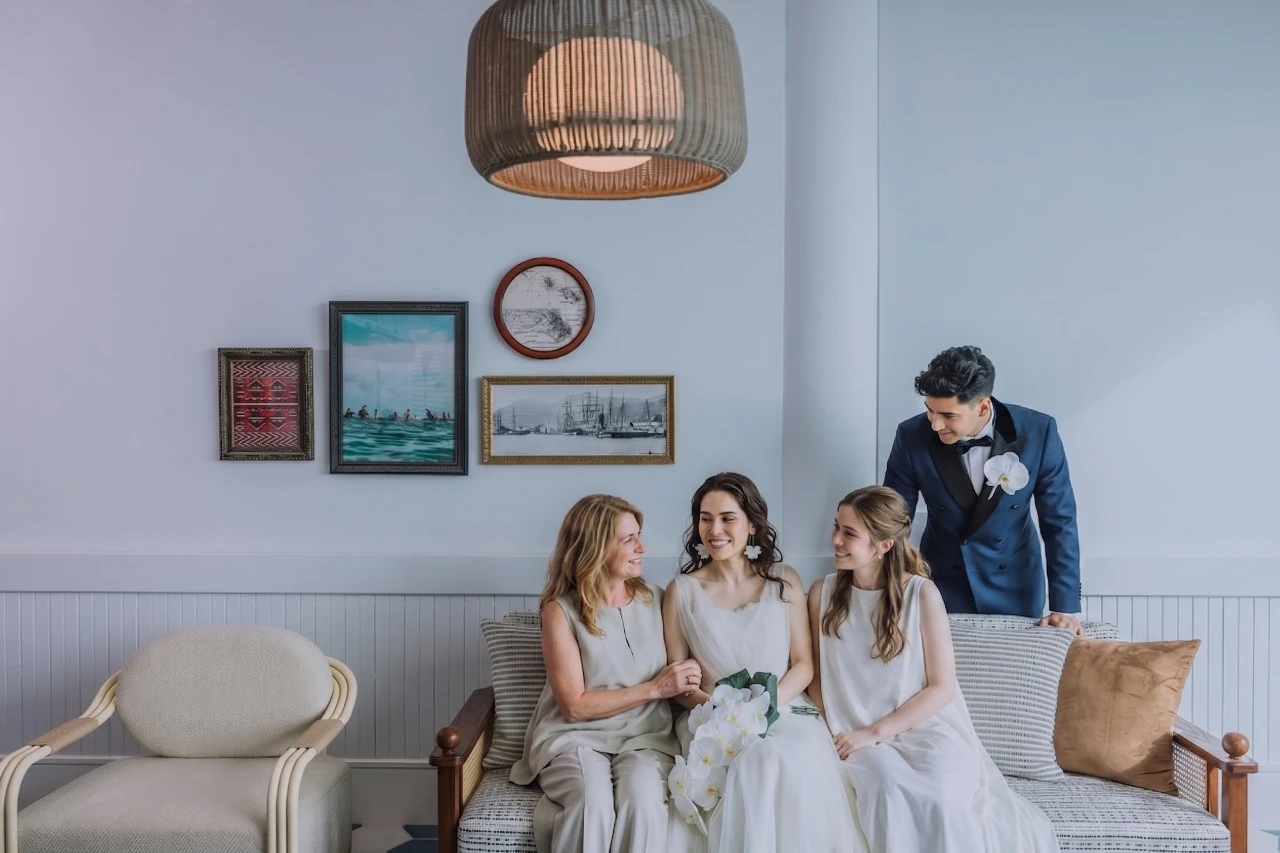 A family smiles warmly while sitting on a modern tropical couch inside a beach-house style lounge. The bride, wearing a white gown and holding a bouquet of white orchids, sits in the center flanked by two women in light-colored attire, while a groom in a navy tuxedo with a white orchid boutonniere stands behind the couch looking down at them. Above them hangs a large woven wicker pendant light against a soft gray wall adorned with framed ocean and coastal artwork.