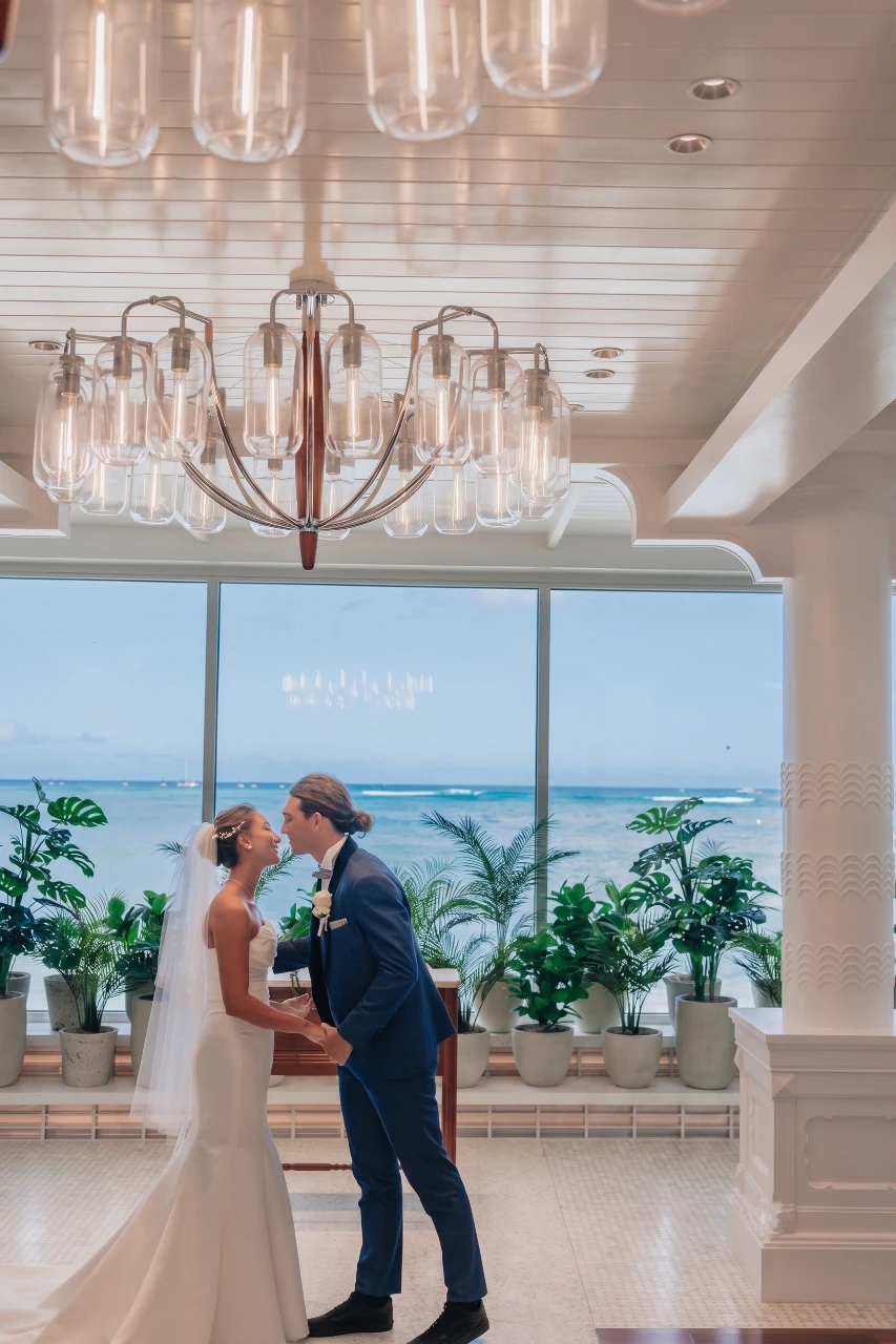 A bride in a fitted white wedding gown and veil holds hands with a groom in a blue suit as they share an intimate smile inside Kahawai Chapel. Behind them, large floor-to-ceiling glass windows reveal a panoramic view of the sparkling blue Pacific Ocean, bordered by lush tropical potted plants under a modern, glowing glass chandelier.