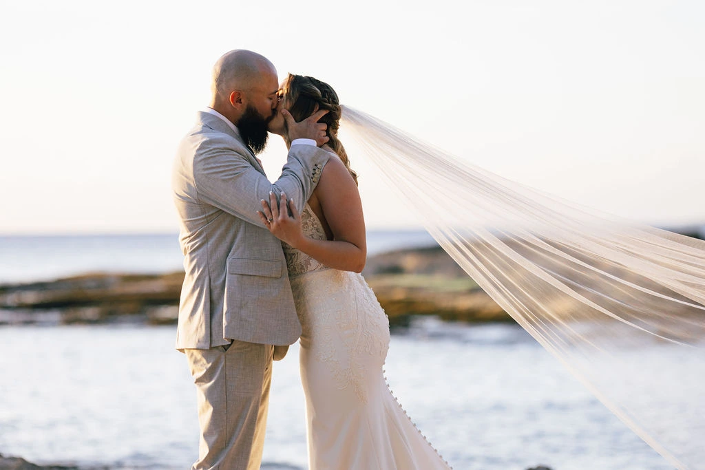 A bride in an embroidered white gown and long flowing veil shares an embrace and kiss with the groom in a light grey suit against a scenic ocean backdrop