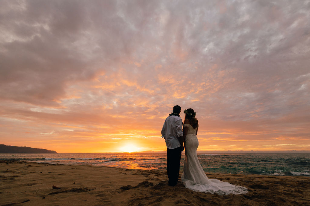 Bride in lace gown and groom in maile lei holding hands during a sunset beach wedding at Papailoa Beach on Oahu's North Shore.