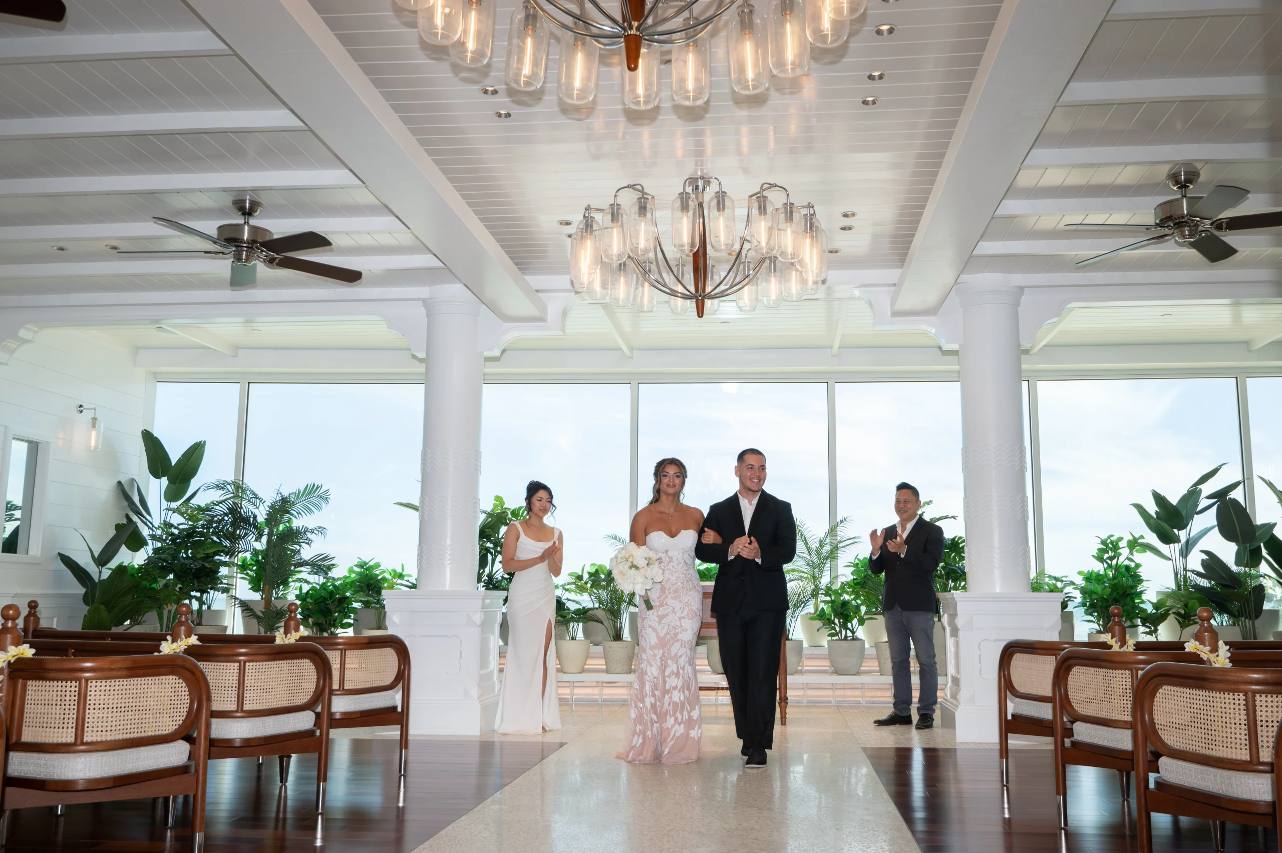 A bride in a strapless lace wedding dress holding a white floral bouquet walks down the aisle arm-in-arm with her groom in a black suit inside a bright, sunlit chapel with tropical plants, grand chandeliers, and guests applauding.