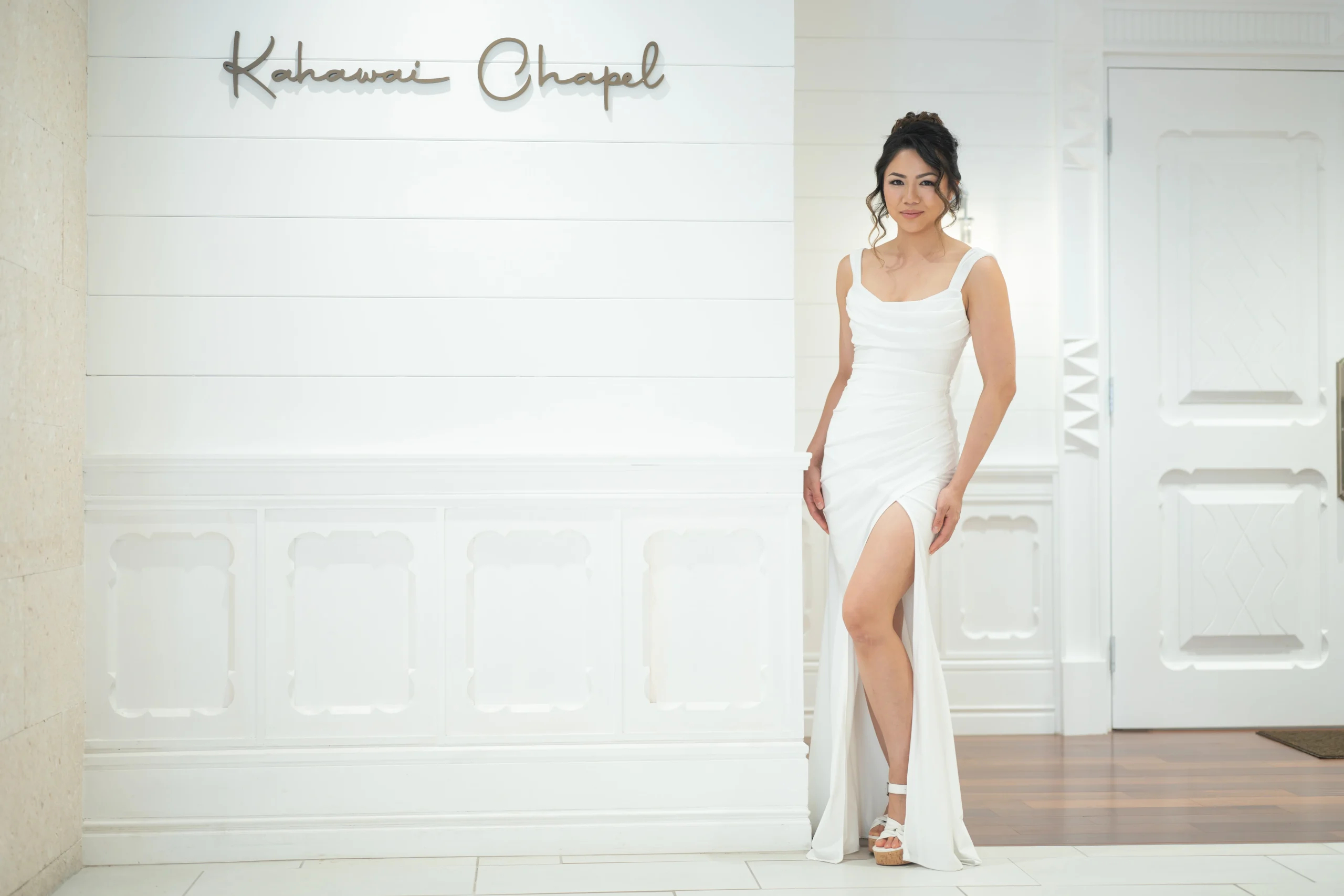 A bride in a sleek white sleeveless wedding gown with a thigh-high slit poses by the white shiplap entrance of Kahawai Chapel