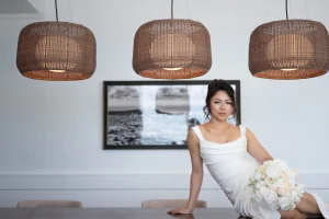 A bride in a sleek white sleeveless wedding dress reclines across a dark wooden table holding a white floral bouquet beneath three large woven pendant lights