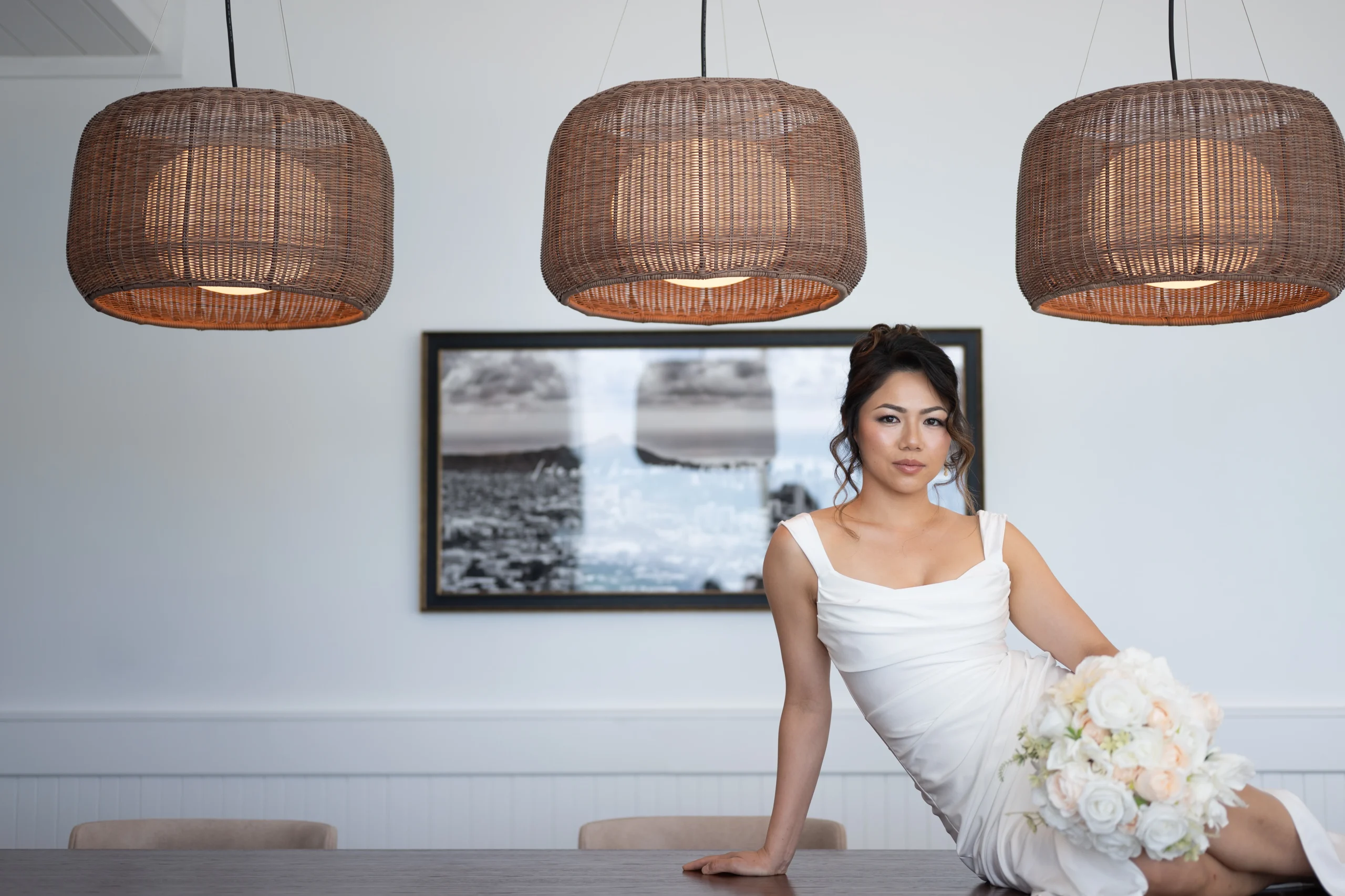 A bride in a sleek white sleeveless wedding dress reclines across a dark wooden table holding a white floral bouquet beneath three large woven pendant lights