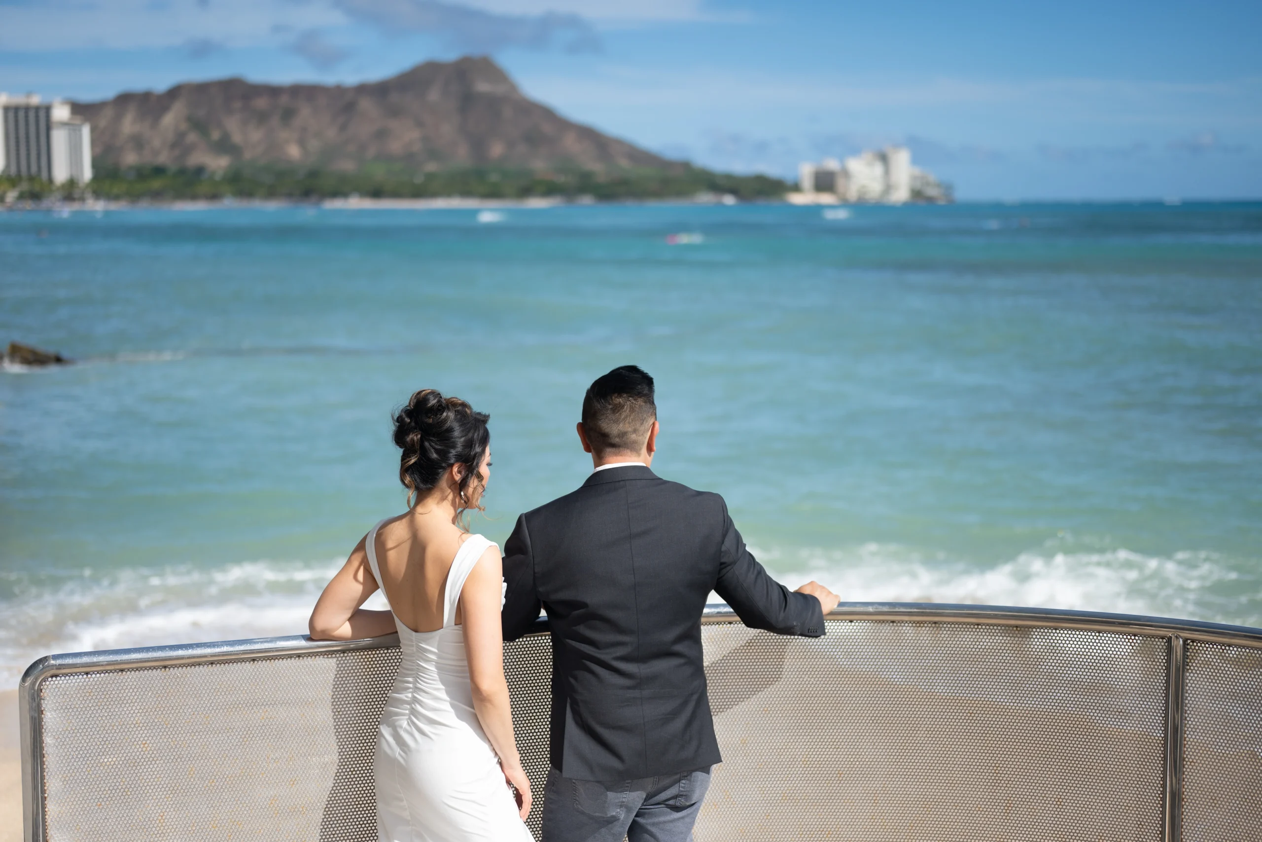 Rear view of a bride in an open-back white dress and groom in a dark suit jacket leaning against a railing, gazing out at Diamond Head and the turquoise ocean in Waikiki