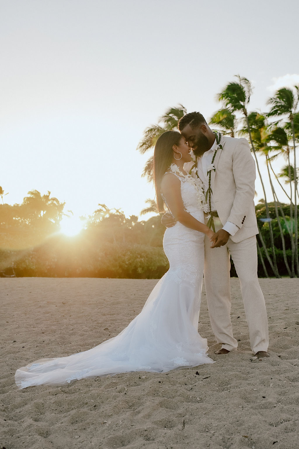 Bride in mermaid lace gown and groom in beige linen suit embracing at golden hour, one of the best sunset wedding locations on Oahu.