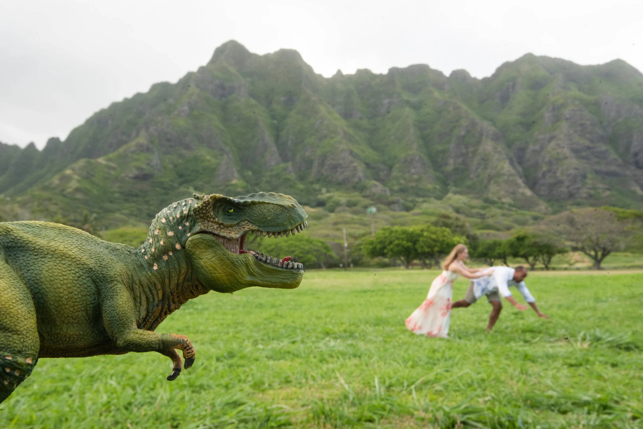 Bride and groom running playfully from a toy T-Rex during Kualoa Regional Park weddings backed by the Koʻolau Mountains