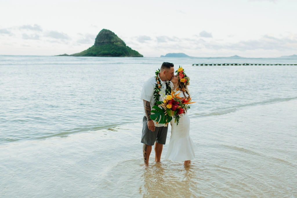 Bride and groom kissing in the calm ocean shallows during Kualoa Regional Park weddings facing Mokoliʻi Island